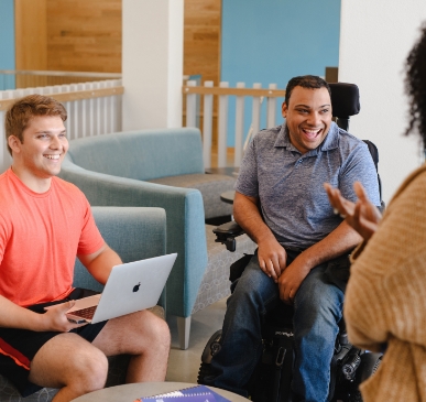 A student with a laptop and another student in a wheelchair smile and laugh while engaging in conversation with a professor in a classroom full of natural light