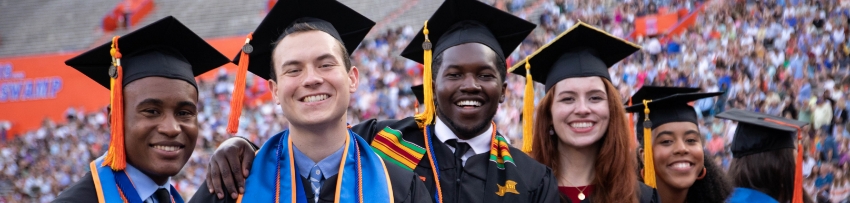 Six alumni of diverse backgrounds smile widely at the camera at their graduation while wearing caps, gowns and academic achievement cords and stoles
