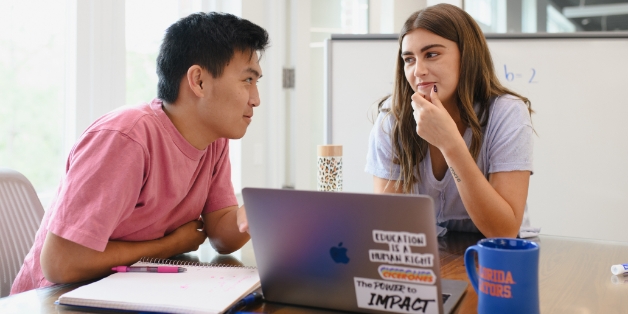 Two students lightly smile at one another and converse in front of a laptop while the student on the left points at the laptop screen and the student on the right gestures thoughtfully