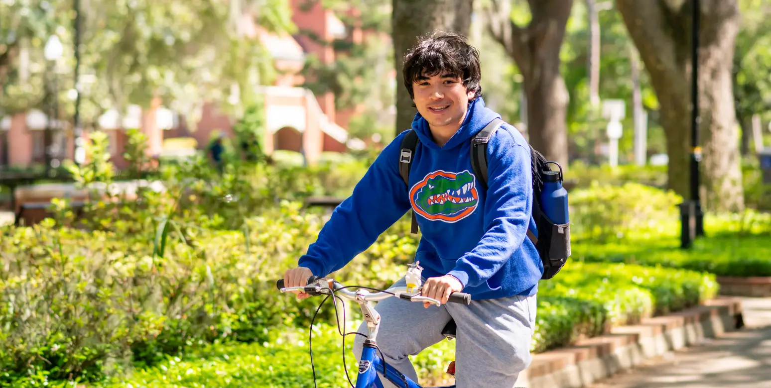 A young adult smiles while cycling across the green University of Florida campus