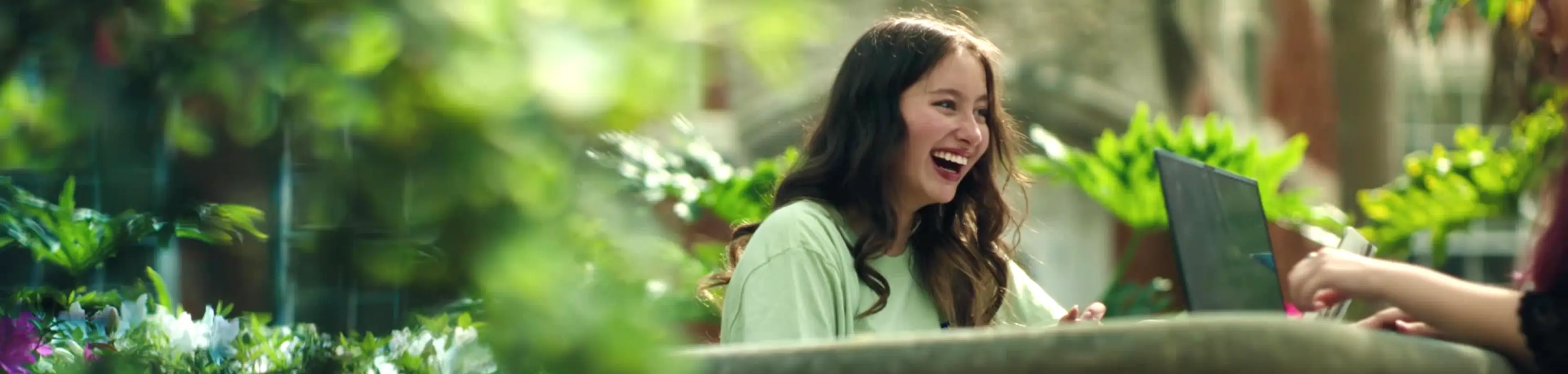 A young adult smiles and laughes with another student outside at a UF picnic table with their laptops