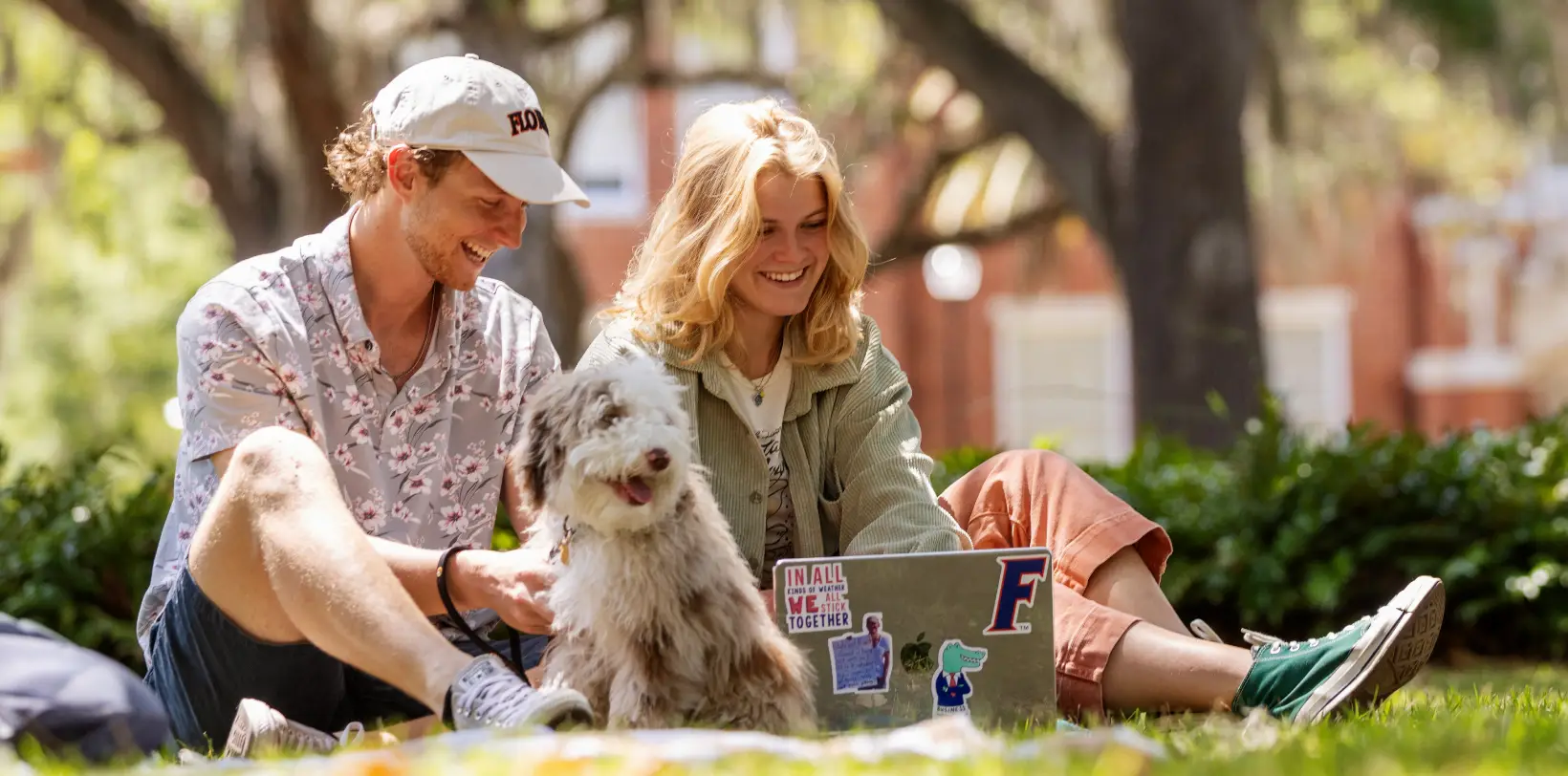 Two students sit with a small dog and laptop on the Plaza of Americas lawn at the University of Florida